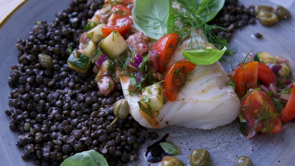 Close-up of a gourmet fish fillet with lentils, tomato, and fresh herbs on a plate.